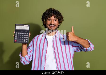 Porträt des Buchhalters arabischer Angestellter in gestreiftem lässigem Hemd, das den Daumen nach oben zeigt und den Rechner isoliert auf khakifarbenem Hintergrund hält Stockfoto