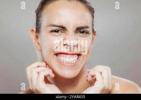 Nahaufnahme einer fröhlichen jungen Frau, die ihre perlweißen Zähne zeigt, während sie ihre Fäuste spielerisch vor einem neutralen Hintergrund zusammendrückt. Stockfoto