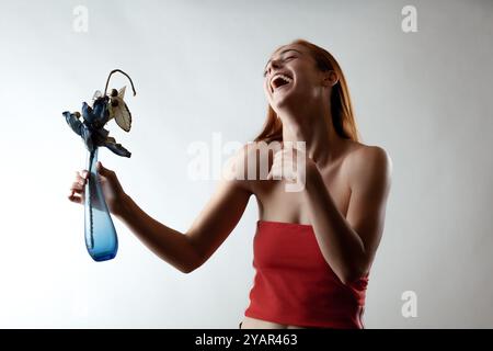 Eine verspielte, fröhliche junge Frau in einem roten Bandeau lacht herzhaft, während sie eine skurrile Mottenskulptur hält. Das Bild fängt einen Moment des echten Glücks ein Stockfoto