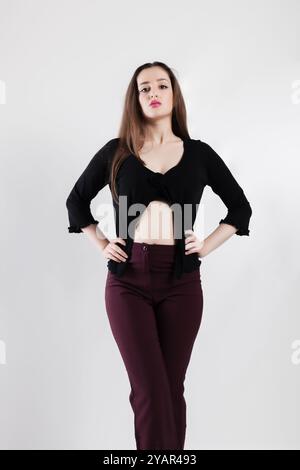 Eine junge Frau posiert selbstbewusst in einem schwarzen Crop Top und einer burgunderroten Hose vor einem schlichten weißen Hintergrund. Stockfoto