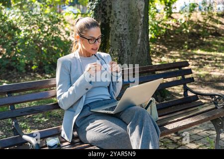Junge erfolgreiche Geschäftsfrau, die Videoanrufe auf einem Laptop macht, während sie auf der Bank im Park sitzt. Geschäftsfrau arbeitet ferngesteuert, spricht mit Stockfoto
