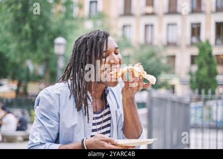 Porträt eines afroamerikanischen Mannes, der durch die Stadt spaziert, während er ein Stück Pizza, Fast Food-Konzept beißt. Stockfoto