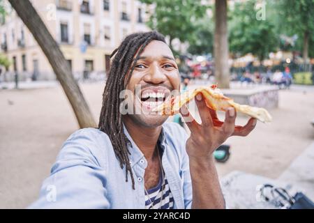 Ein aufgeregter Mann, der Selfie macht, während er Pizza isst. Lachender Mann mit Dreadlocks, der Spaß beim Abendessen hat. Stockfoto