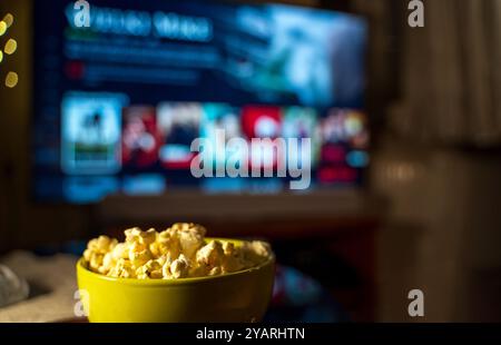 Eine Schale Popcorn im Vordergrund mit einem Fernseher im Hintergrund, der eine Streaming-Service-Schnittstelle zeigt. Stockfoto