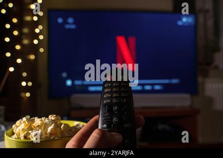 Krakau, Polen - 15. Oktober 2024: Nahaufnahme einer Hand mit Fernbedienung, mit einem Fernseher im Hintergrund, der das Netflix-Logo zeigt. Eine Schüssel p Stockfoto