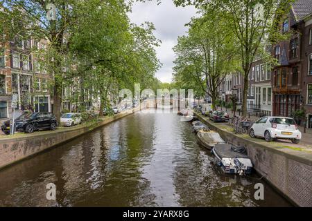 Der belaubte und baumgesäumte Reguliersgracht-Kanal im Herzen von Amsterdam, Niederlande, vom 28. Mai 2024 mit geparkten Autos und Booten Stockfoto