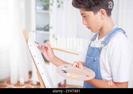 Ein kleiner Junge malt ein Bild mit einem Pinsel und einer Palette. Er trägt einen blauen Overall und ein weißes Hemd. Der Junge konzentriert sich auf seine Malerei und t Stockfoto