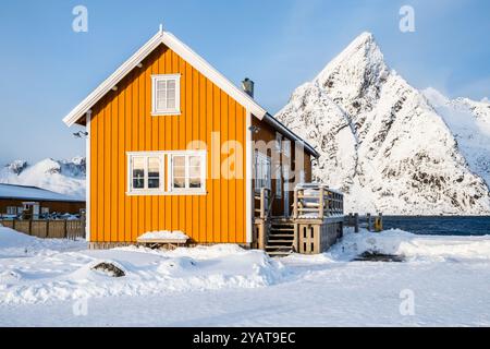 Traditionelles gelbes Rorbu-Haus und der Berg Olstind im Fischerdorf Sakrisoy auf den Lofoten-Inseln in Norwegen. Winterlandschaft mit schneebedeckten Bergen Stockfoto