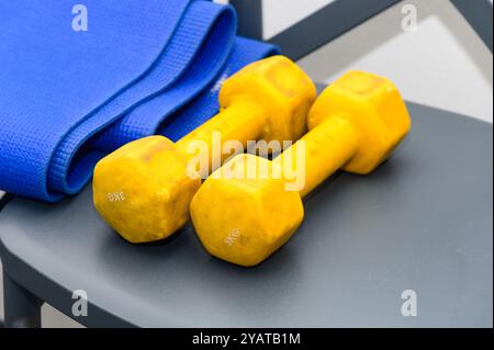 Zwei leuchtend gelbe Kurzhanteln sitzen auf einem eleganten grauen Stuhl, begleitet von einer ordentlich gerollten blauen Yogamatte, die eine einladende Workout-Umgebung schaffen. Stockfoto