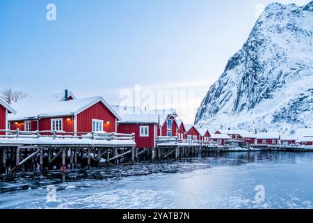 Reines Dorf mit traditionellen Rrorbu-Häusern auf den Lofoten-Inseln im Winter. Skandinavische Landschaft mit roten hölzernen Fischerhütten auf gefrorenem Meerwasser Stockfoto