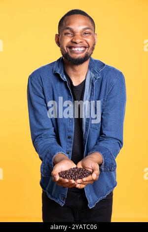 afroamerikaner fröhliche Person posiert mit schwarzen gerösteten Kaffeebohnen vor gelbem Hintergrund, Koffeinliebhaber. Junger Erwachsener, der arabische Americano-Kaffeesamen empfiehlt. Stockfoto