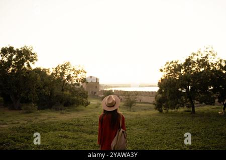 Frau mit Hut und orangefarbener Jacke steht auf einem Feld und blickt während des Sonnenuntergangs auf eine alte Festung, umgeben von der Natur, und genießt friedliche Momente während des Sonnenuntergangs Stockfoto