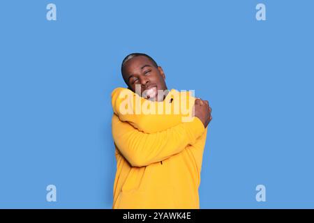 Ein fröhlicher Schwarzer mit einem knallgelben Hoodie umarmt sich mit einem großen Lächeln vor einem leuchtend blauen Hintergrund, der Glück und Kommen ausstrahlt Stockfoto