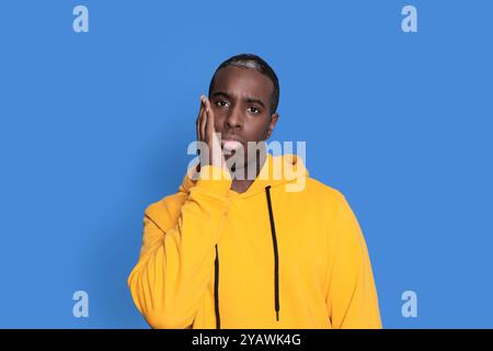 Ein junger Schwarzer mit einem knallgelben Hoodie hält seine Wange und sieht besinnlich vor einem schlichten blauen Hintergrund aus, was Introspektion vermittelt. Stockfoto