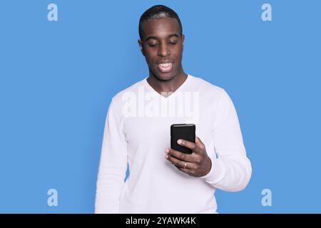 Ein fröhlicher Schwarzer in einem weißen Pullover schaut glücklich auf sein Smartphone, steht vor einem schlichten blauen Hintergrund und strahlt eine lässige und entspannte Atmosphäre aus. Stockfoto