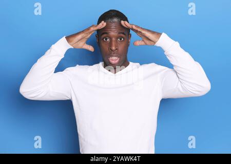 Ein schwarzer Mann in einem weißen Pullover sieht schockiert aus, seine Hände auf dem Kopf, die Augen weit offen, vor blauem Hintergrund, was eine starke Überraschung oder Unzufriedenheit ausdrückt Stockfoto