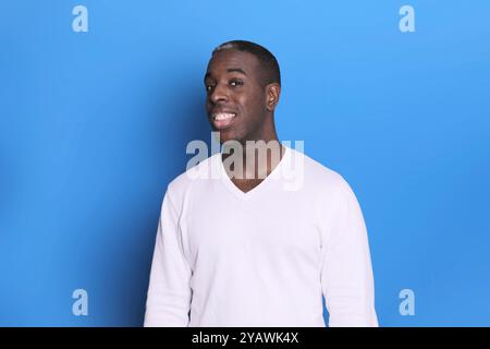 Ein selbstbewusster Schwarzer in einem weißen Pullover steht vor einem schlichten blauen Hintergrund und blickt mit einem freundlichen, offenen Ausdruck direkt in die Kamera. Stockfoto