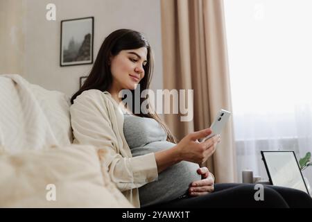 Schöne lächelnde schwangere Frau mit Babybauch Handy benutzen, online einkaufen, im Internet surfen, Podcast schauen, während sie zu Hause auf dem Sofa sitzt. Arzt-Videoanruf-Arzttermin Stockfoto