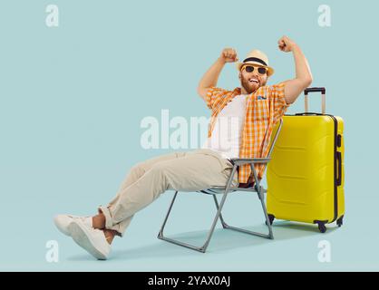 Fröhlicher, lustiger Reisender, der triumphierend sitzt und die Arme im Sieg in der Nähe eines gelben Koffers erhoeht. Stockfoto