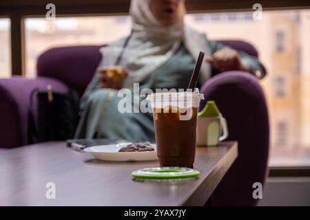 Gekühlter Eiskaffee, serviert in einer Plastiktasse mit schwarzem Strohhalm, begleitet von einem Schokoladenkeks auf einem weißen Teller. Perfekt für eine gemütliche Kaffeepause Stockfoto