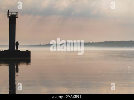 Silhouette eines einsamen Mannes, der neben einem kleinen Turm am Ende eines Piers im Morgennebel steht. Stockfoto