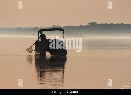 Silhouette eines einsamen Mannes, der einen Fisch auf seinem Pontonboot im Morgennebel fängt. Stockfoto