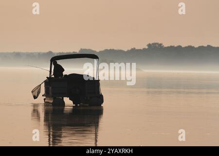 Silhouette eines einsamen Mannes, der einen Fisch auf seinem Pontonboot im Morgennebel fängt. Stockfoto