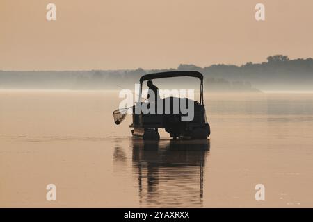 Silhouette eines einsamen Mannes, der einen Fisch auf seinem Pontonboot im Morgennebel fängt. Stockfoto