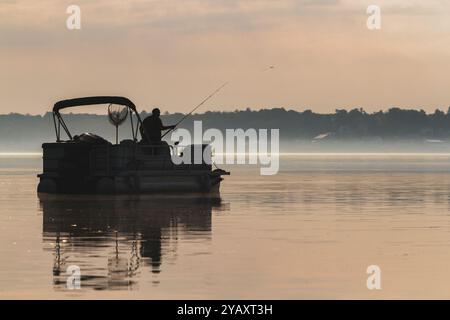 Silhouette eines einsamen Mannes, der seine Rute wirft, während er im Morgennebel auf seinem Pontonboot fischt. Stockfoto