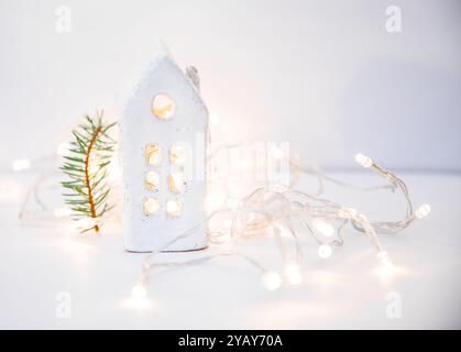 Minimalistisches Weihnachtsdekor aus kleinem Keramikhaus und weiße Neujahrslampen auf weißem Hintergrund. Stockfoto