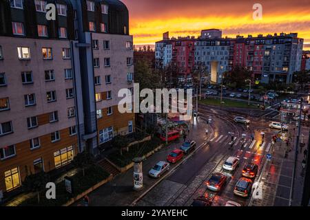 Eine lebhafte Stadtszene mit einer geschäftigen Kreuzung bei Sonnenuntergang. Autos und Fußgänger bewegen sich durch nasse Straßen, während die Gebäude mit Innenbeleuchtung leuchten Stockfoto