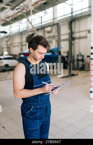 Vertikale Aufnahme eines fokussierten Automechanikers, der das Auto mit Clipboard im Garagendienst überprüft. Ernsthafter Techniker, der das Fahrzeug in der Garage repariert Stockfoto