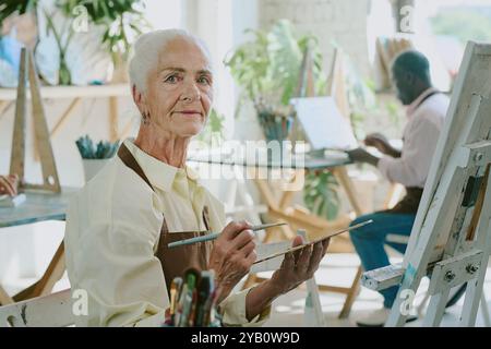 Ältere Frau, die Palette mit Ölfarben und Pinsel hält, während sie im Zeichenstudio in die Kamera schaut Stockfoto