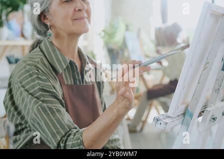 Mittlere Nahaufnahme der Hand einer älteren kaukasischen Frau, die einen Pinsel hält, während sie Striche auf weißer Leinwand macht Stockfoto