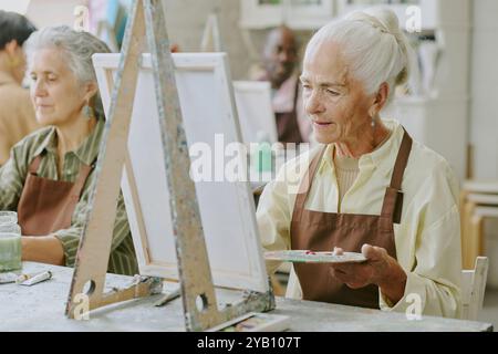 Weißhaarige ältere Frau, die Holzpalette mit gemischten Farben hält, während sie auf Leinwand streichelt Stockfoto
