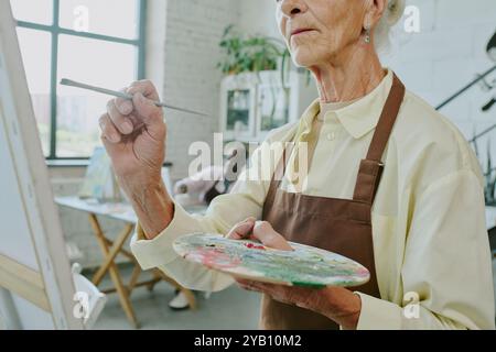 Niedrigwinkelaufnahme einer nicht erkennbaren Seniorin, die schmutzige Palette und Pinsel hält, während sie Kunstwerke auf Leinwand erstellt Stockfoto