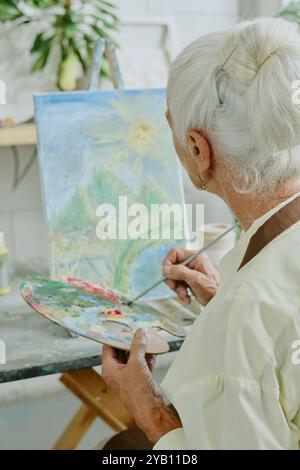 Rückansicht Aufnahme einer nicht erkennbaren Seniorin, die Ölfarben auf Holzpalette mischt und dabei die Landschaft auf Leinwand bewundert Stockfoto