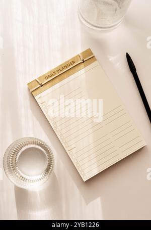 Flacher, minimalistischer Tagesplaner mit einfachem Design, Kontrollkästchen und Linien für Aufgaben. Helle Oberfläche neben einem Glasbecher, wodurch eine saubere Oberfläche entsteht Stockfoto