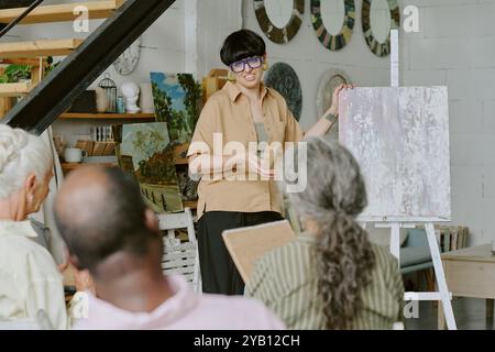 Rückansicht einer Gruppe von Personen, die einer jungen Lehrerin zuhören, die vor der Schülergruppe im Studio Zeichentipps erklärt Stockfoto