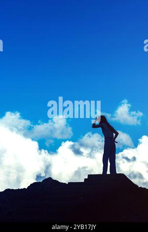 Die junge Frau steht zuversichtlich auf einem felsigen Gipfel, die Silhouette vor einem leuchtenden Himmel voller Wolken. Sie blickt in die Ferne Stockfoto