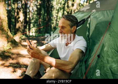 Reifer Mann sitzt in einem Zelt und benutzt ein Smartphone. Senior Mann mit Smartphone im Wald. Stockfoto