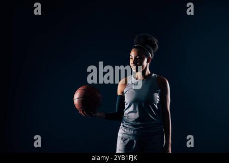 Junge Frau in Mannschaftssportbekleidung, die einen Basketball vor einem schwarzen Hintergrund hält Junge Frau in Mannschaftssportbekleidung, die einen Basketball vor einem schwarzen Hintergrund hält Stockfoto