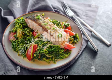 Saftig geröstetes Lachsfilet mit knuspriger Haut auf Gemüse aus Rahmspinat, Zwiebeln und Tomaten, gesundes Fischgericht mit Besteck auf grauer Serviette und Stockfoto