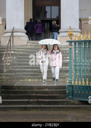 Ein paar junge weibliche Touristen mit einem Regenschirm verlassen das University Fitzwilliam Museum in Cambridge, England, an einem regnerischen Tag. Stockfoto