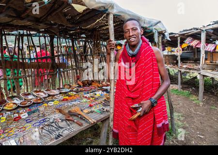 Männliches Mitglied des Masai-Stammes in traditioneller roter Kleidung. Neben einem Souvenirstand, Masai Mara, Kenia, Afrika Stockfoto