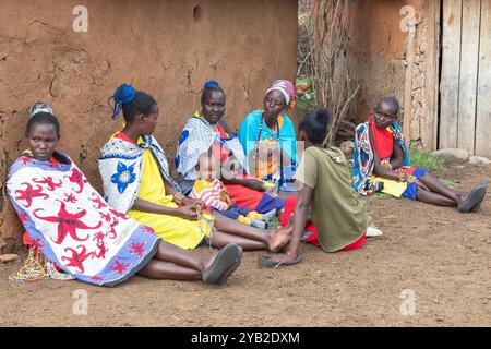 Gruppe von Masai-Frauen, die traditionelle Kostüme tragen, sitzen und plaudern vor einem traditionellen Haus aus Kuhmist und -Schlamm, Masai Village, Stockfoto