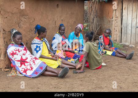 Gruppe von Masai-Frauen, die traditionelle Kostüme tragen, sitzen und plaudern vor einem traditionellen Haus aus Kuhmist und -Schlamm, Masai Village, Stockfoto