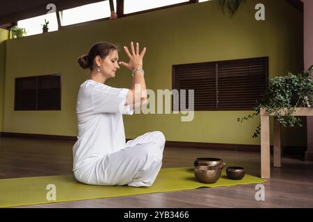 Sportliche Frau, die Sukhasana-Übungen macht, in bequemer Lotusposition auf der Matte im Zimmer oder Yoga-Studio sitzt, Namaste-Geste zeigt, Training in weiß Stockfoto