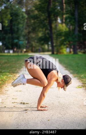 Frau, die Handstand macht, ein Balancierendes Asana namens Bakasana im Yoga, Kranhosen, in lässiger Kleidung auf einem Wanderweg in einem Park üben Stockfoto