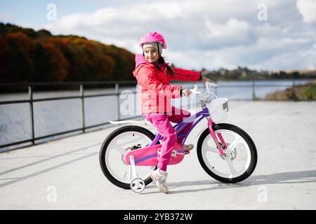 Ein junges Mädchen mit rosa Outfit und Helm macht an einem sonnigen Tag eine malerische Radtour am See. Die lebhaften Farben und die natürliche Umgebung erinnern an einen Stockfoto